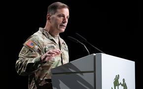 A military officer in camouflage uniform speaks at a podium with dual microphones against a black background. 
