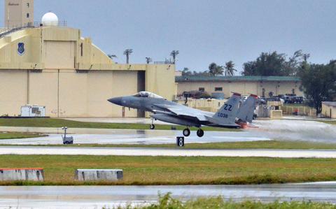 A military fighter jet takes off from a runway with an aircraft hangar and control tower in the background.