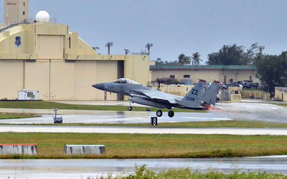A military fighter jet takes off from a runway with an aircraft hangar and control tower in the background.