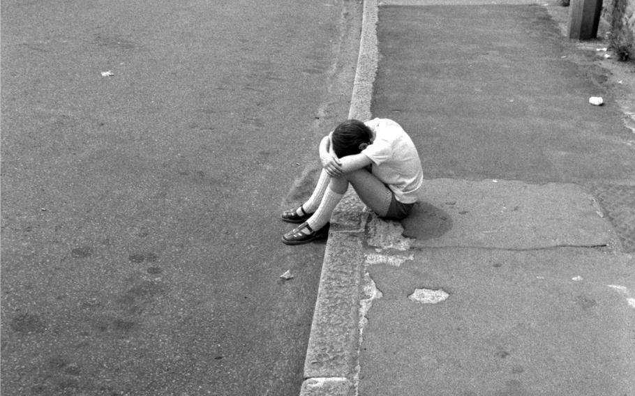 A black-and-white, 1970s-era image of a boy in t-shirt and shorts sitting on a curbside with his head down on his arms.