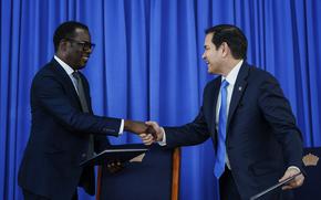 Marco Rubio grins while shaking hands with his Guyanese counterpart, against a blue curtain.
