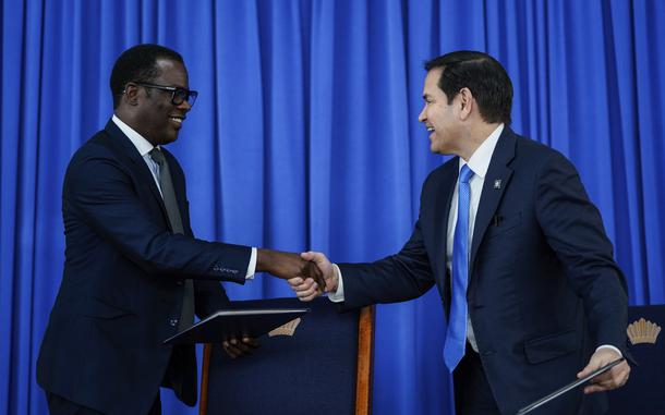 Marco Rubio grins while shaking hands with his Guyanese counterpart, against a blue curtain.