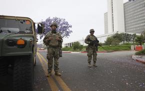 Two soldiers in camouflage combat gear, one holding a machine gun, stand next to an armored vehicle in a street in front of a multi-story office building.