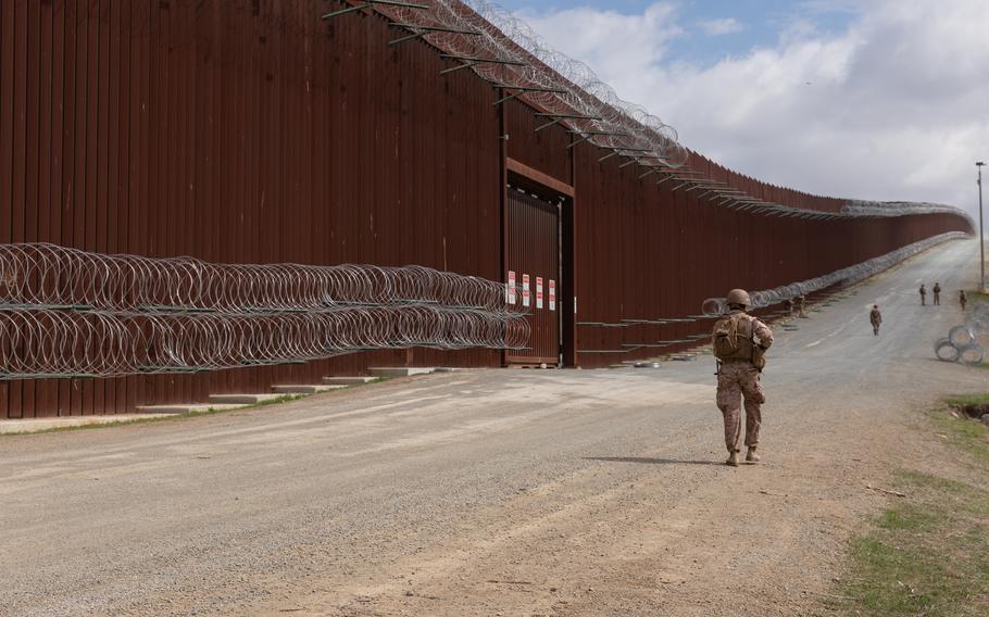Marines walk on a dirt road along a wall with barbed wire.