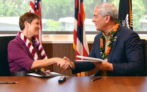 A woman in a purple shirt and Hawaiian lei shakes hands with a man in a blue blazer and Hawaiian lei while seated at a table, with U.S. and Hawaii flags behind them in front of a window.