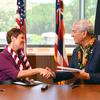 A woman in a purple shirt and Hawaiian lei shakes hands with a man in a blue blazer and Hawaiian lei while seated at a table, with U.S. and Hawaii flags behind them in front of a window.