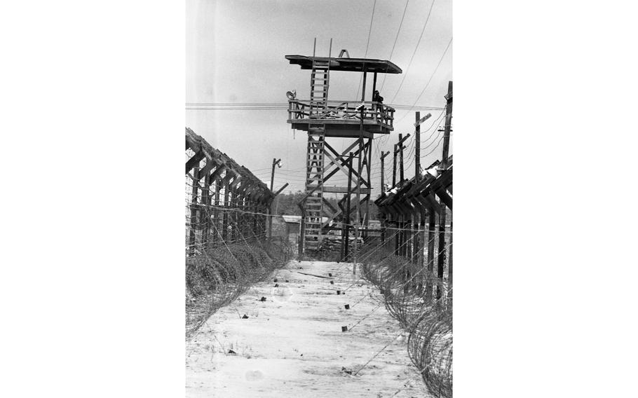 An Army of the Republic of Vietnam Military Police (ARVN QC) guard stands atop a guard tower at Bien Hoa POW camp, overlooking the multiple barbed wire fences that surround the camp. 