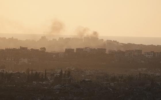 Smoke rises over a hazy city skyline with destroyed buildings in the foreground.