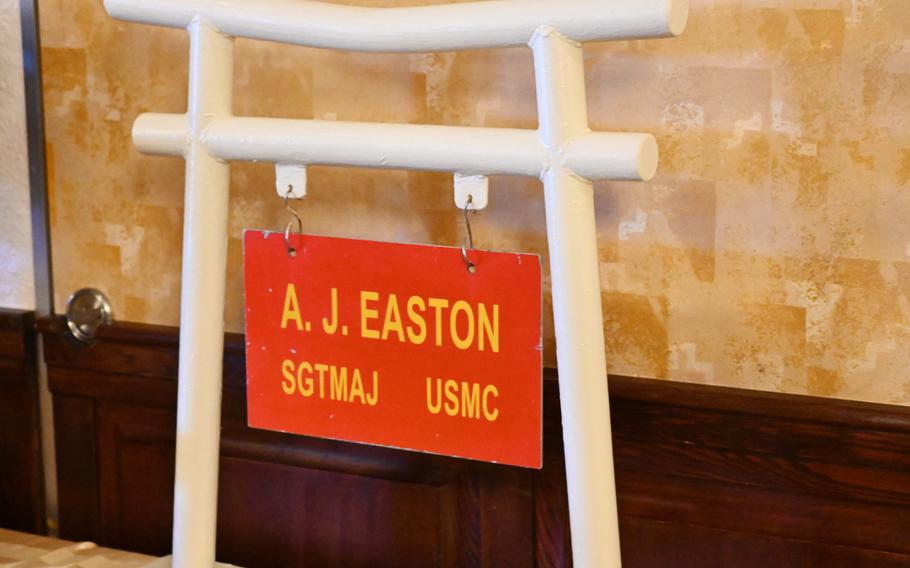 A white Japanese torii gate with a red sign reading “A.J. Easton SGTMAJ USMC” hanging underneath stands in front of a beige wall.