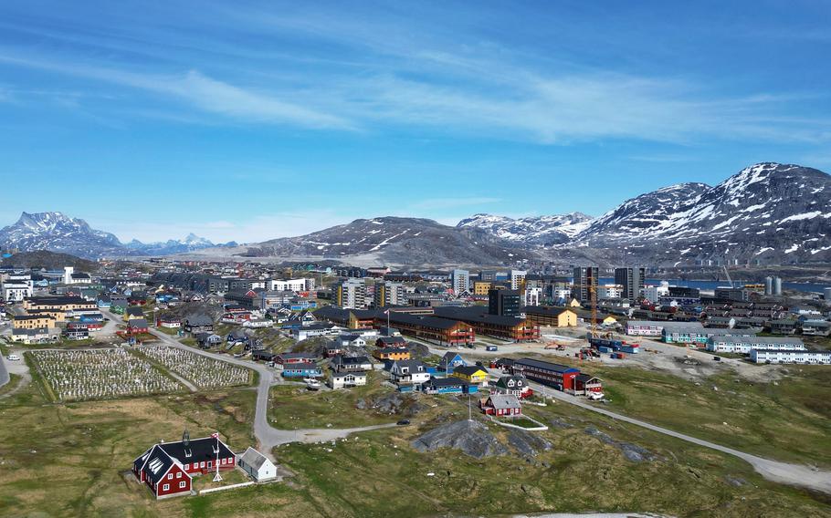 An aerial view of homes and buildings in the cityscape of Nuuk, Greenland, with mountains and blue sky in the background.
