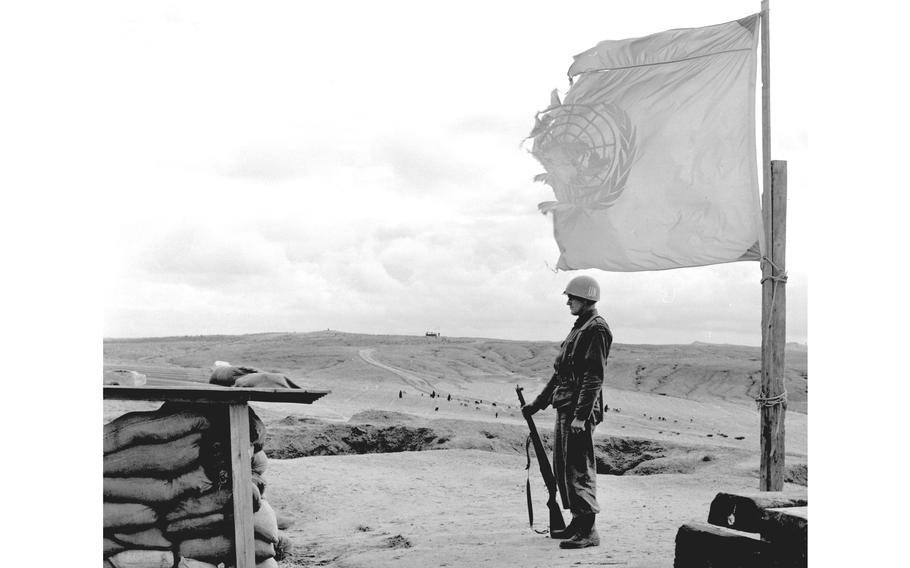 A black and white photo of service member standing next to a torn U.N. flag, his rifle resting on the ground.
