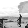 A black and white photo of service member standing next to a torn U.N. flag, his rifle resting on the ground.