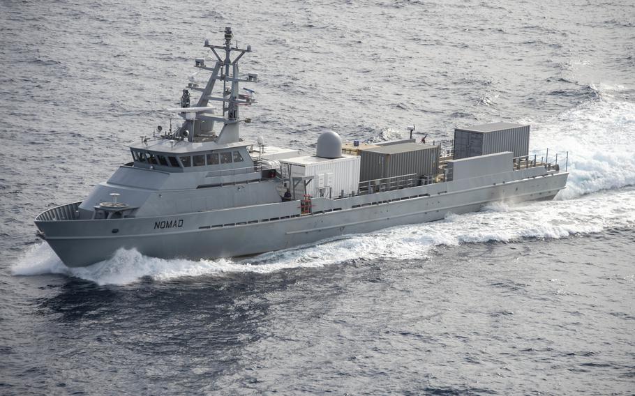 A gray military patrol vessel named “NOMAD” cuts through choppy ocean waters, creating white wake spray at its bow. The ship features a prominent superstructure with radar equipment, communication arrays and cargo area with shipping containers on the rear deck. 