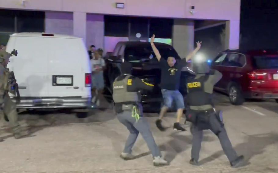 A blurry image of a man holding up his arms as two law enforcement officers seem to aim weapons at him.