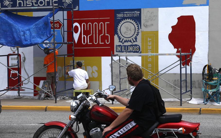 A motorist watches owner Daniel Ricketts, left, and muralist Jerome Lamke with St. Louis Sign & Mural, hand-paint a Route 66 mural on a building at the southeast corner of East Vandalia and South Main streets on July 25 in Edwardsville, Ill. The mural is one of 11 highlighting Route 66 in southern Illinois towns. 