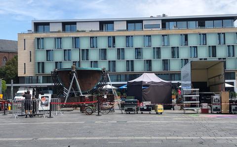 A  production setup in an urban plaza featuring large professional lighting equipment with curved reflectors mounted on metal stands, surrounded by various production gear including equipment cases, tents and barriers. The scene is set against a modern multi-story building with distinctive green-tinted glass panels and light-colored facade. 