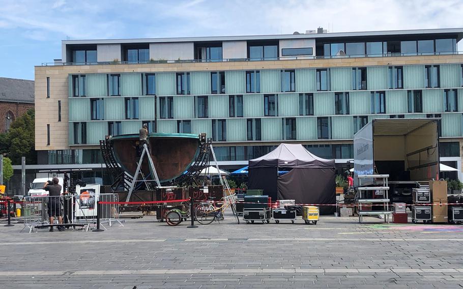 A production setup in an urban plaza featuring large professional lighting equipment with curved reflectors mounted on metal stands, surrounded by various production gear including equipment cases, tents and barriers. The scene is set against a modern multi-story building with distinctive green-tinted glass panels and light-colored facade. 