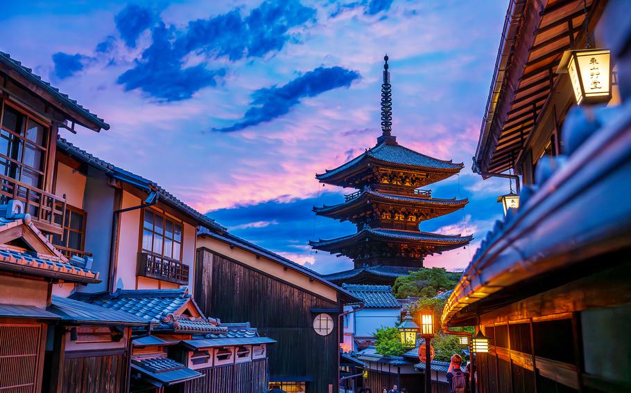 A traditional Japanese pagoda is seen between the eaves of other buildings as blue and purple clouds form around it at sunset.