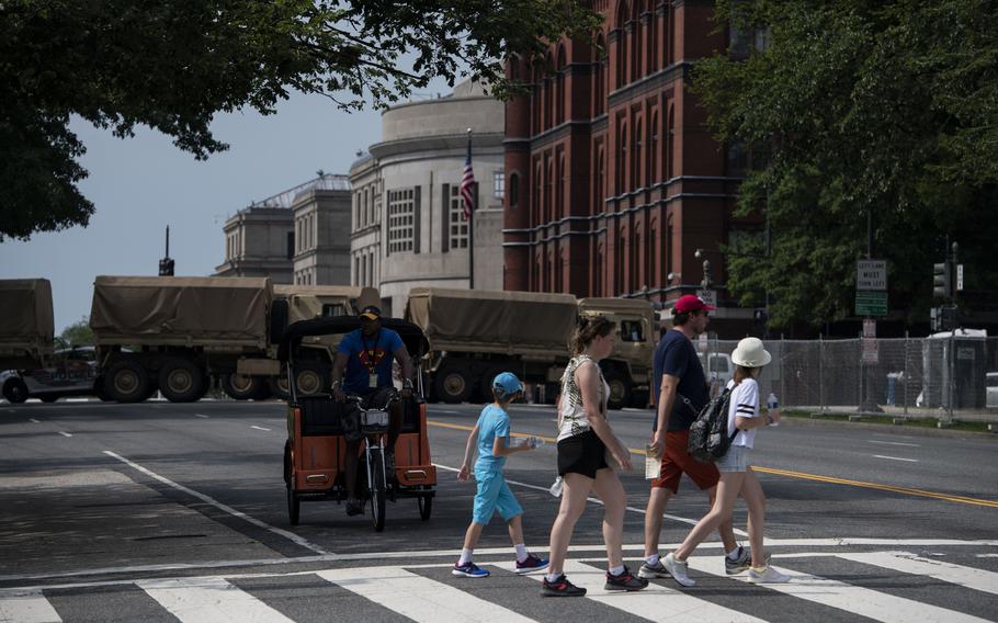 Pedestrians walk across a street. Trucks blocking the street are in the background.