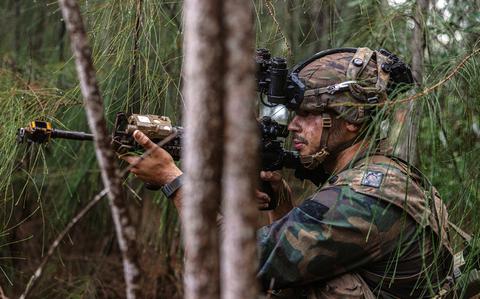 Seen through trees, a soldier in profile wearing a helmet with binoculars aims his rifle.