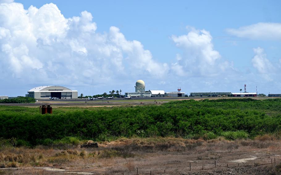 An airfield complex with a hangar and observation tower are seen from a distance, with a cloudy sky in the background and a grassy field in the foreground.