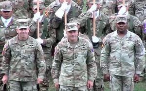 Three Army officers in camouflage uniforms and hats stand in a row at attention in front of other soldiers in camouflage uniforms and hats during a ceremony.