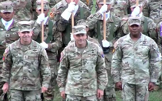 Three Army officers in camouflage uniforms and hats stand in a row at attention in front of other soldiers in camouflage uniforms and hats during a ceremony.