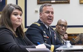 A military officer in dress uniform sits at a table in front of a microphone next to a woman in black in a congressional hearing room.