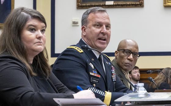 A military officer in dress uniform sits at a table in front of a microphone next to a woman in black in a congressional hearing room.