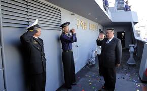 Kim Jon Un, wearing a suit and tie, salutes while standing on the deck of a military ship. Two uniformed naval officers salute him as he passes by. Several other uniformed personnel stand in formation on the upper deck. Confetti is scattered on the floor.