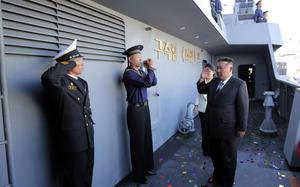 Kim Jon Un, wearing a suit and tie, salutes while standing on the deck of a military ship. Two uniformed naval officers salute him as he passes by. Several other uniformed personnel stand in formation on the upper deck. Confetti is scattered on the floor.