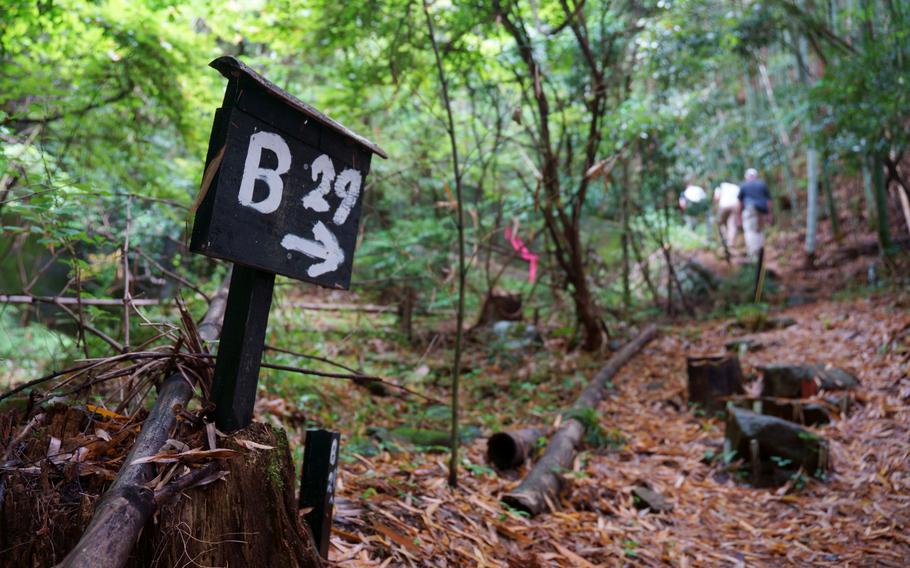 A black wooden marker with “B 29” and an arrow pointing to the right painted in white stands next to a forest trail with hikers in the background.