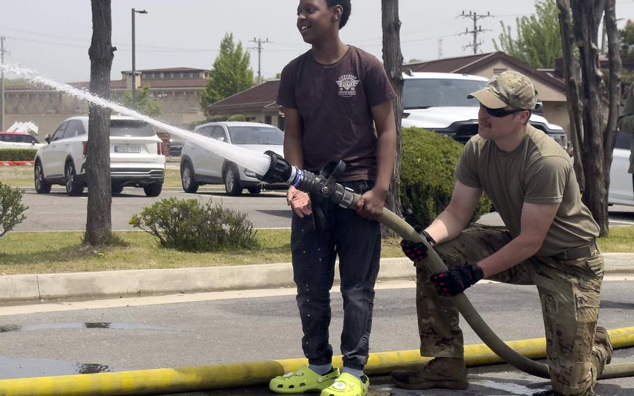 A smiling young boy holds a large fire hose spraying water with the assistance of a kneeling airman. They are on a wet street surrounded by vehicles and trees.