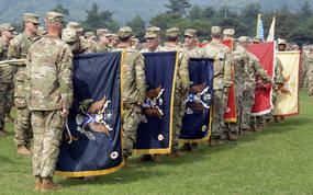 Pairs of soldiers in camouflage uniforms stand facing each other with flags held between them during a transfer ceremony on a grassy field.