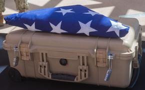 A folded American flag with white stars visible on blue fabric rests on top of a tan military-style hard case. The flag is ceremonially folded in the traditional triangular manner typically used for military honors and funeral services. 