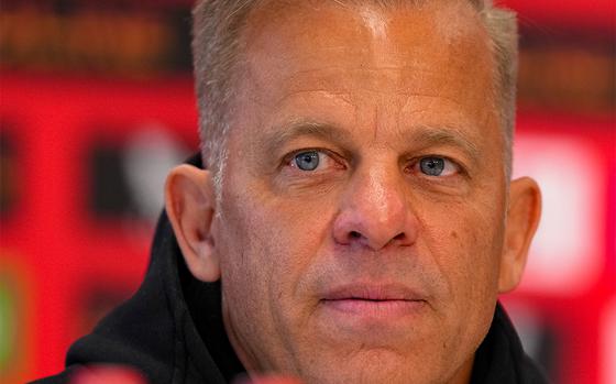 A close-up of a man with short, light blond hair and blue eyes, wearing a black hoodie, speaking at a press conference with a red and black backdrop behind him.