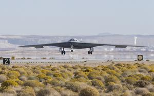 A stealth bomber, seen from a distance, lands on a runway with scrub land in the foreground.