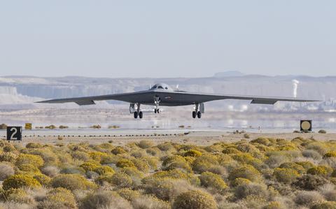 A stealth bomber, seen from a distance, lands on a runway with scrub land in the foreground.