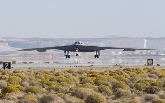 A stealth bomber, seen from a distance, lands on a runway with scrub land in the foreground.