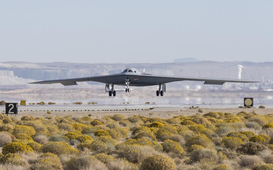 A stealth bomber, seen from a distance, lands on a runway with scrub land in the foreground.