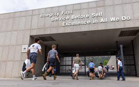 Military personnel and cadets walk through a concrete archway or tunnel entrance to a military academy. Large white text on the building reads “Integrity First, Service Before Self, Excellence in All We Do.” 