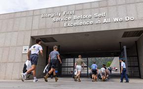 Military personnel and cadets walk through a concrete archway or tunnel entrance to a military academy. Large white text on the building reads “Integrity First, Service Before Self, Excellence in All We Do.” 