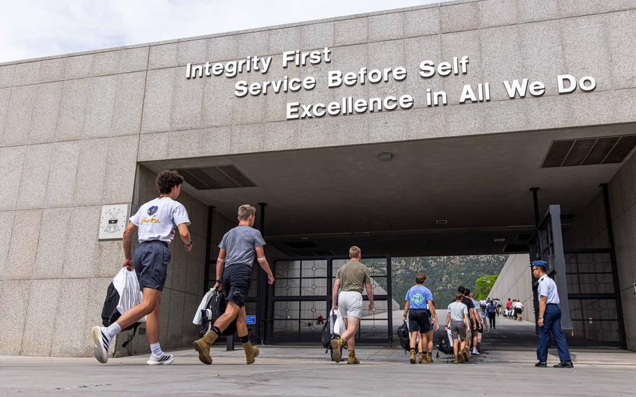 Military personnel and cadets walk through a concrete archway or tunnel entrance to a military academy. Large white text on the building reads “Integrity First, Service Before Self, Excellence in All We Do.” 