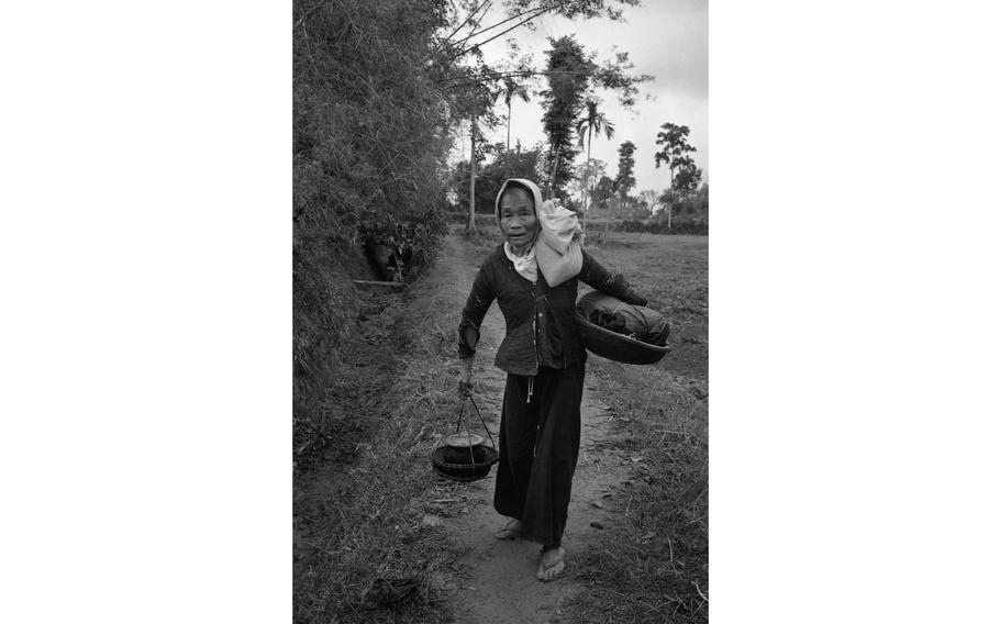 A vertical black and white photo of a woman walking down a dirt pat while carrying containers.