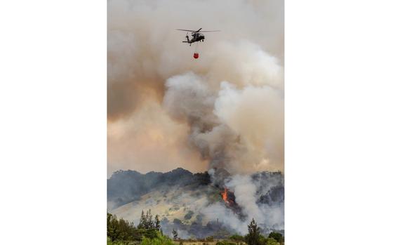 A large plume of smoke billows into the sky from a fire in a forested area as a helicopter hovers above to drop water from a red basket.