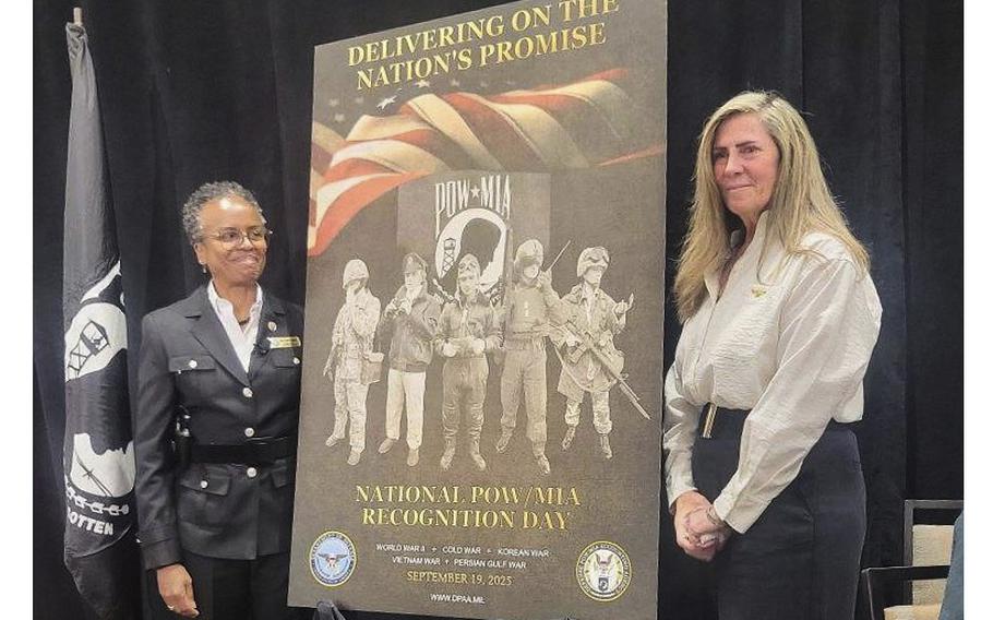 Two women stand on either side of a large poster of military members.