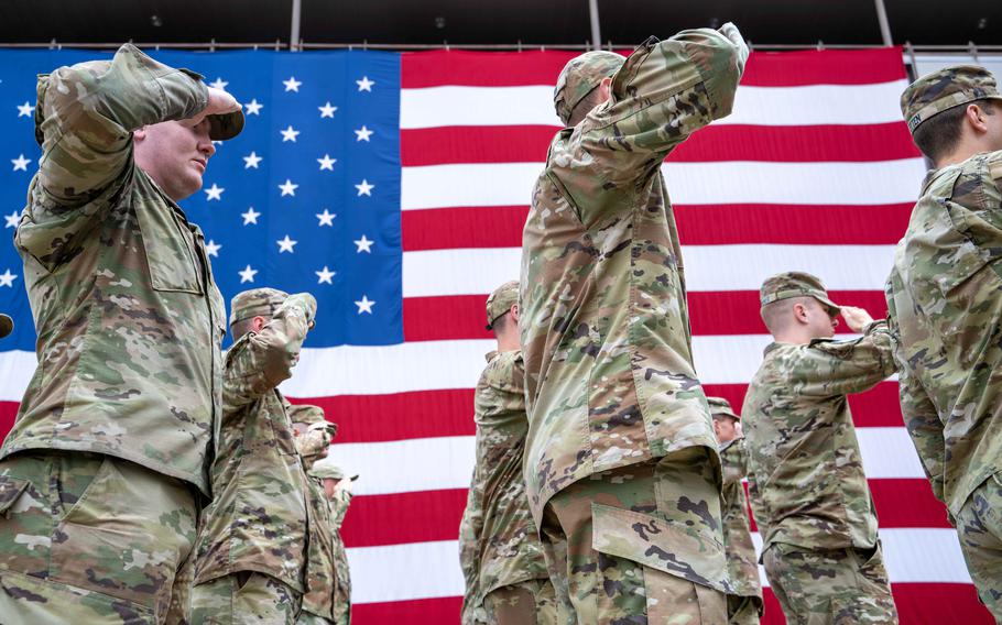Troops in camouflage uniforms and hats salute while standing in formation next to a large U.S. flag.