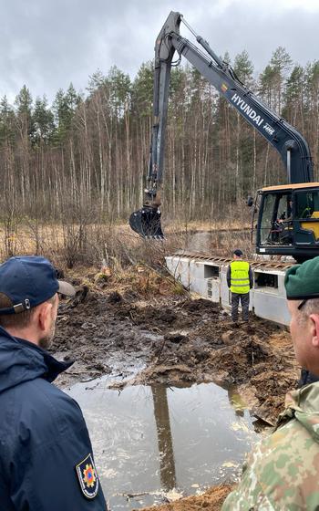 The shoulders of a police officer and Lithuanian service member are seen in the bottom while heavy machinery is seen beyond them.