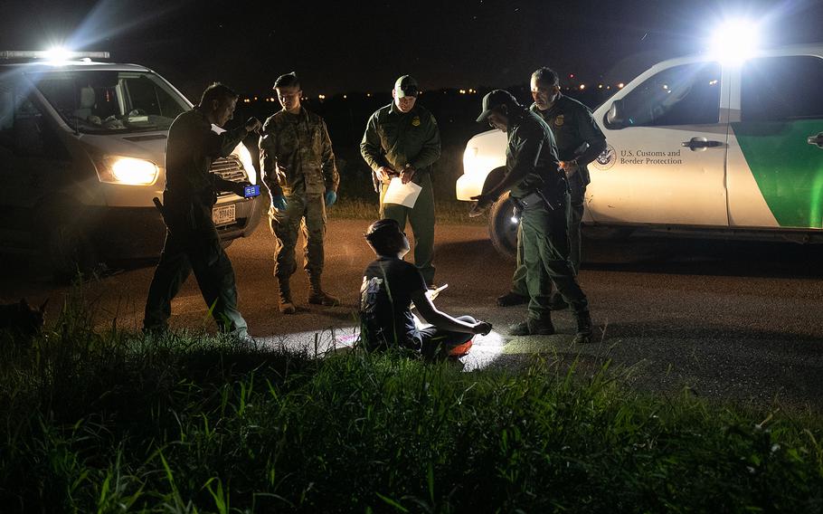 Border Patrol vehicles with bright lights shine on agents and a migrant from China.