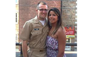 A married couple stand next to each in front of a brick wall, with the man wearing a tan Navy uniform and the woman wearing a floral dress.